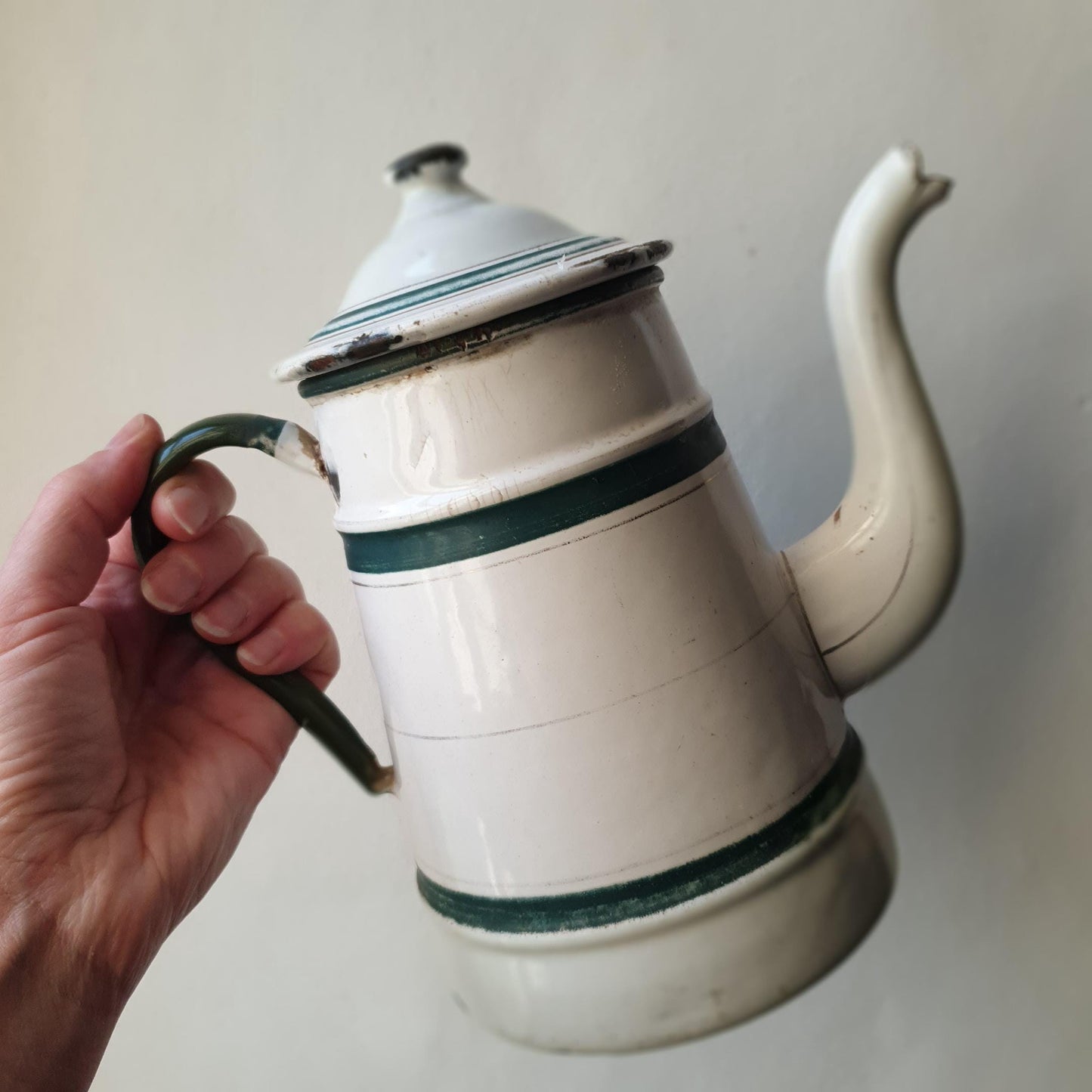 Scale image of a hand holding a vintage French enamel coffee pot from the 1930s shown with its lid, dark green and white enamel with shabby finish
