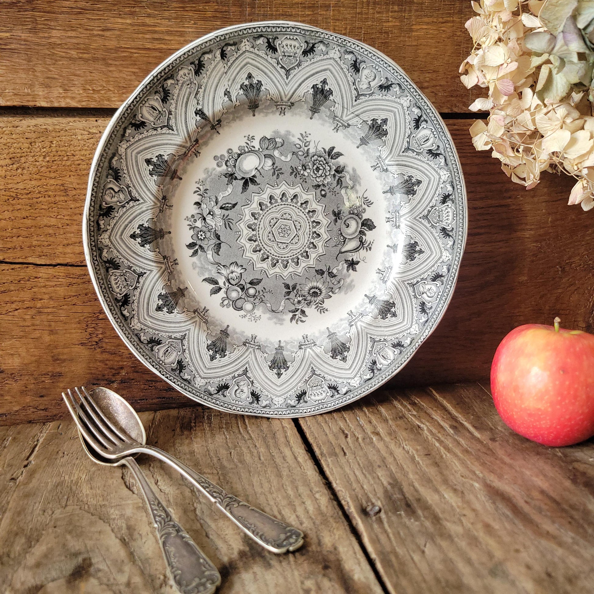 May include: A white and grey patterned plate with a floral and geometric design. The plate is on a wooden surface with a fork, spoon, and a red apple. The plate has a scalloped edge.